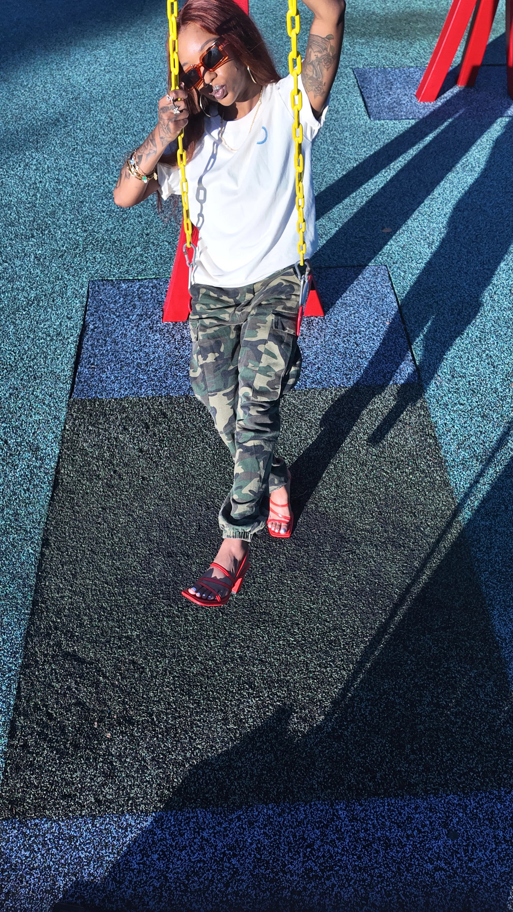 women on red swing with yellow handles wearing bits ad fits unplugged tee, camo cargo pants and orange sandals