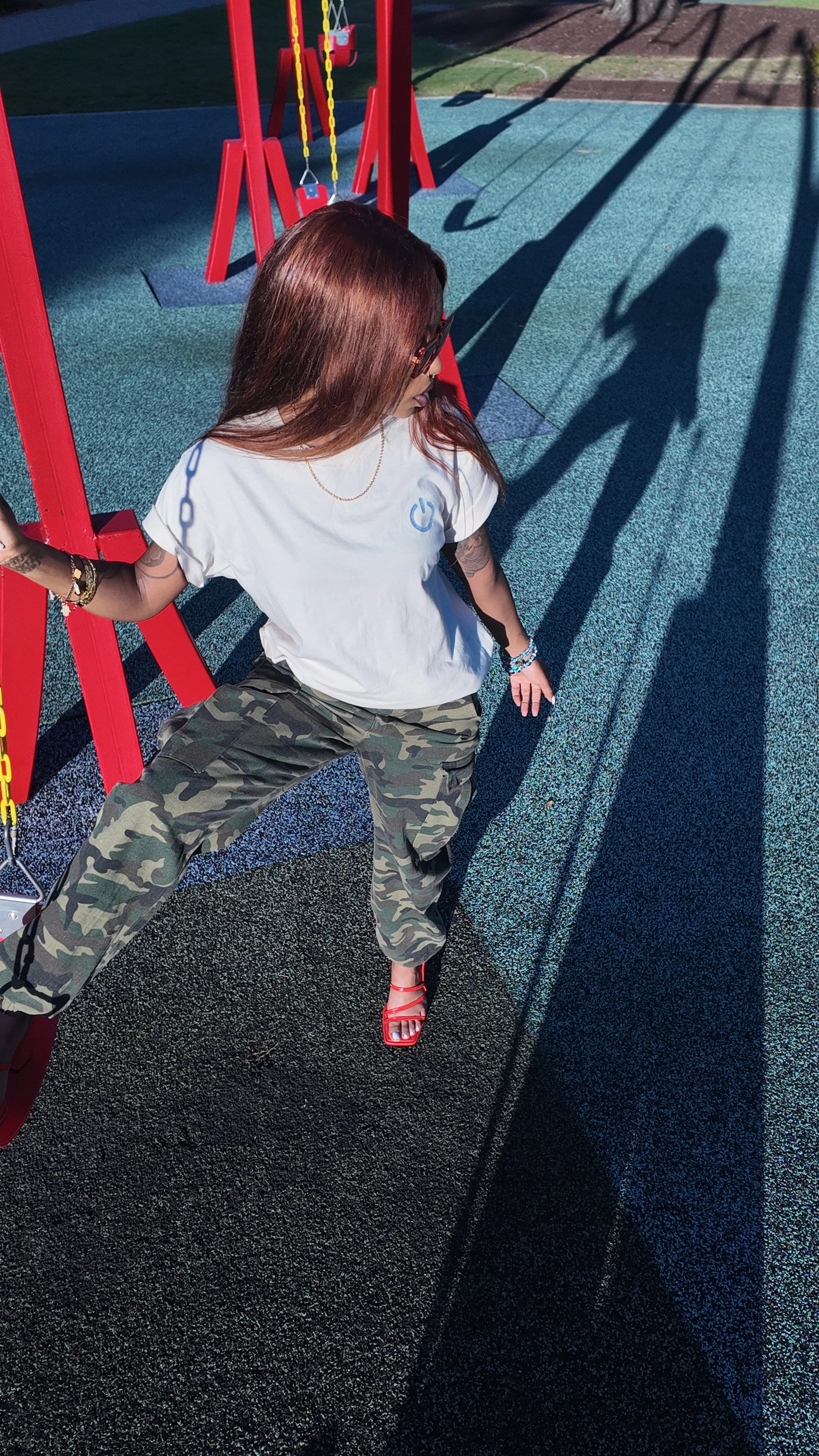 women standing on a playground looking to the left. women is wearing bits and fits unplugged tee, camo cargo pants, and orange heels